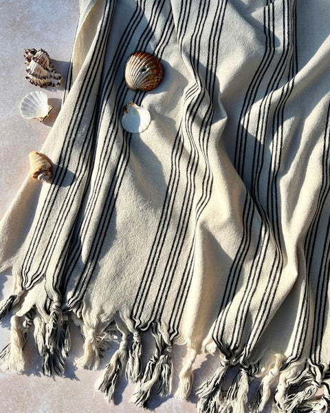 Image of Hydra hammam towel, towel is off-white with fine black stripes at intervals and tassels at the edge. Towel is crumpled in sunlight with a few shells.