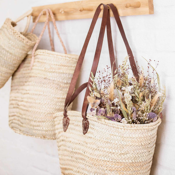 Straw basket bag with chestnut brown leather long and short handles on a hook, filled with dried flowers.