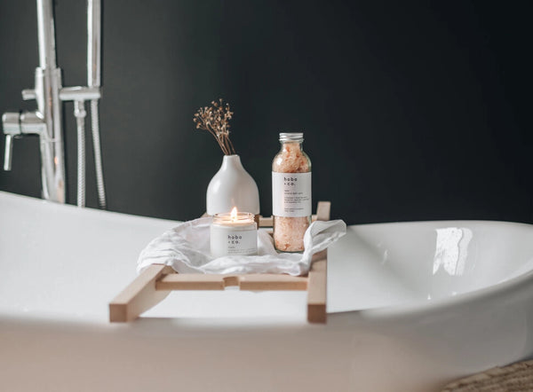 Bath tray with candle, bottle of bath salts and vase over a bath.