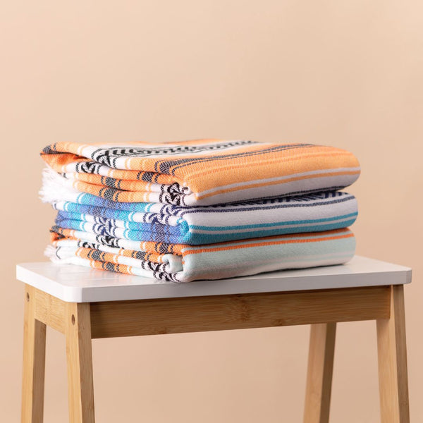 Stack of three Playa hammam towels in orange, blue and aqua, on a wooden stool. From Sand and Salt.