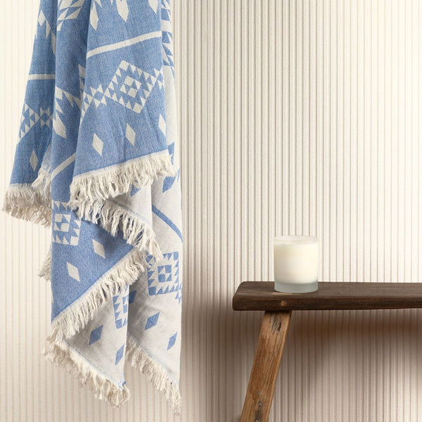 Image of blue patterned hammam towel hanging in a bathroom next to a wooden stool with a white jar candle on it.