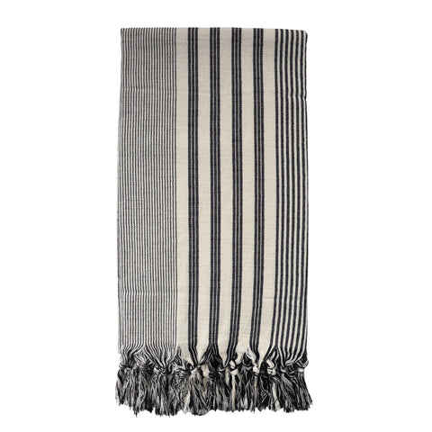 Image is of a folded 100% cotton hammam towel, with a natural base colour and lengthways fine black stripes of varying widths. The towel is finished with soft knotted tassels at both ends.