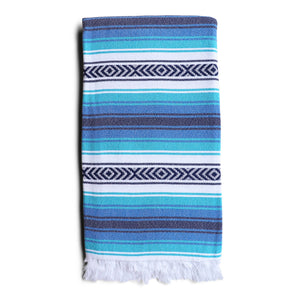 Image is of a folded hammam towel in varying shades of blue and white, with white eyelash fringing at both ends. Sold by Sand and Salt.