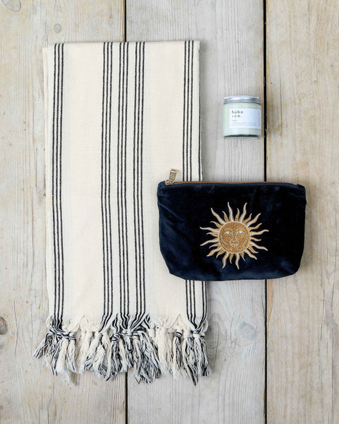 Gift set comprising black and white striped hammam towel, black velvet pouch with gold embroidered sun design and white jar candle on a wooden surface.