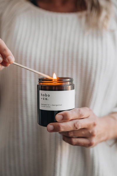Person lighting a jar candle labeled 'hobo co.' balsam fir scent, with a neutral background.