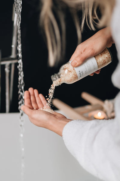 Person pouring bath salts from a bottle into their hand near a bathtub.