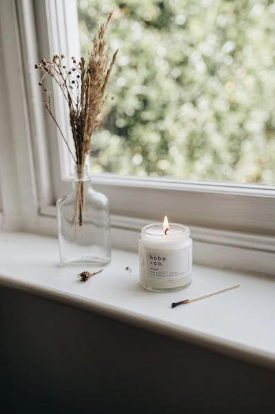 Candle on a windowsill with a vase of dried plants and a blurred outdoor background