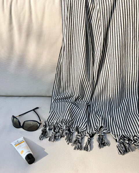 High quality black and white striped hammam towel with tassels, draped over a sunbed, with sunglasses and sunscreen next to it.