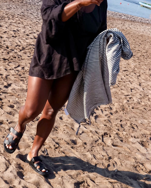 Woman holding a black and white fine striped hammam towel on a sandy beach.