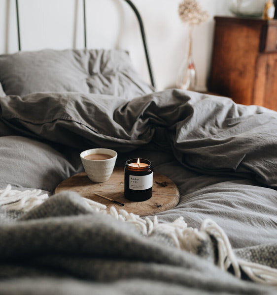 Lit jar candle and mug of tea on a wooden tray on a soft cosy bed with grey bedding.