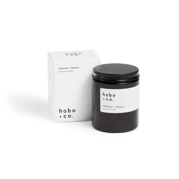 Brown jar candle with white label and black lid next to white packaging on a white background.