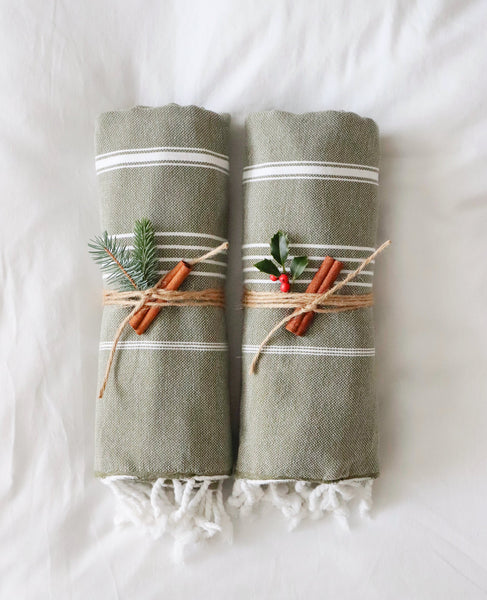 Set of two olive green hammam towels with white stripes, tied with cinnamon sticks and holly leaves on a white background.