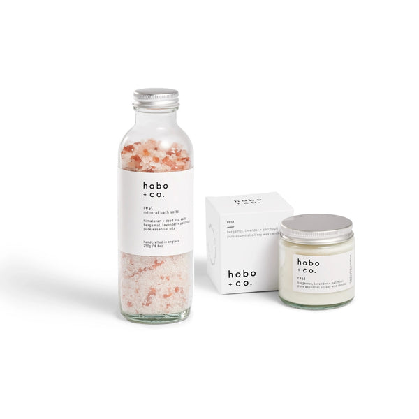 A bath and body gift set from hobo + co. featuring a clear glass bottle filled with pink Himalayan bath salt crystals, a white aromatherapy jar candle with a silver lid, and its white packaging box, all on a clean white background.
