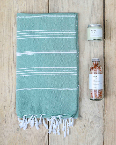 Aqua hammam towel with white stripes and tassels on a wooden surface, accompanied by a jar candle and a bottle of bath salts.