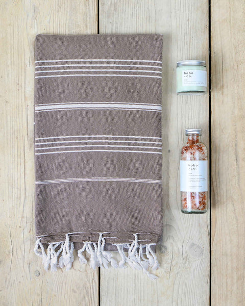 Brown hammam towel with white stripes and tassels on a wooden surface, next to a bottle of bath salts and a jar candle.