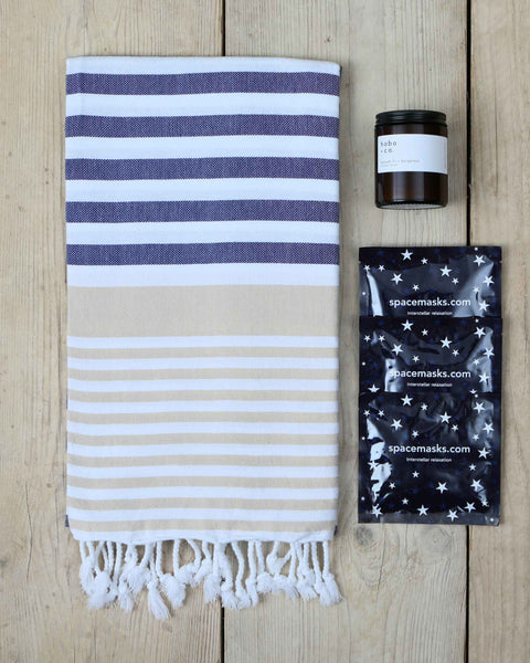 Striped navy, white and beige hammam towel with tassels, a jar candle, and three spacemask warming eyemasks on a wooden surface.