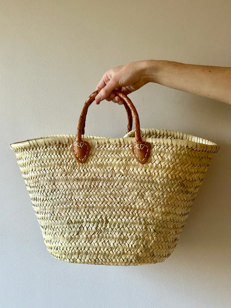 Hand holding a woven basket bag with brown leather handles against a plain background