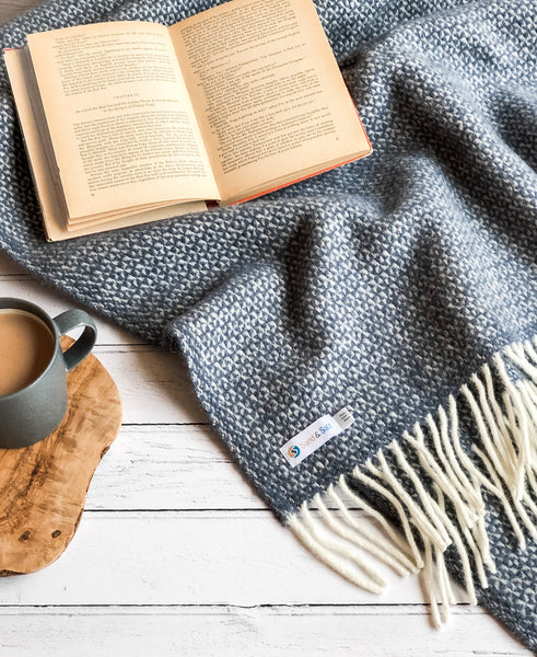 Picture of a welsh wool throw in Slate Blue, folded casually on a table top, with an open book and a mug of tea beside it.
