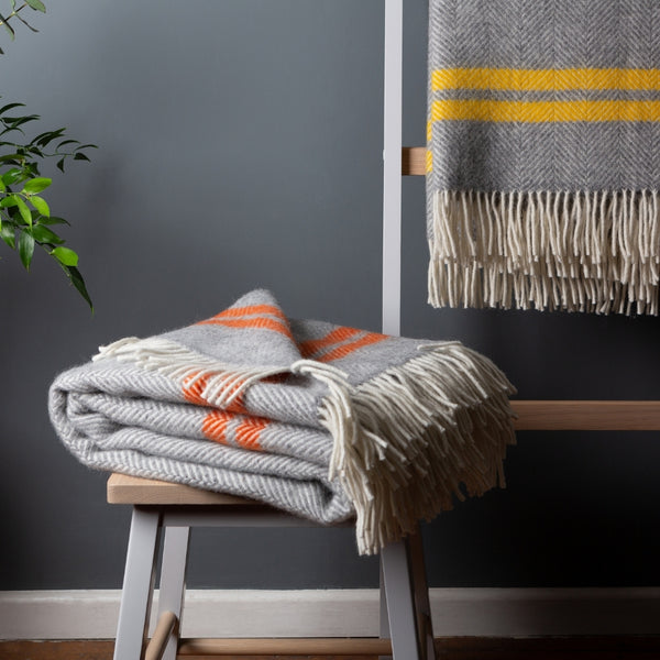 Picture of a light grey wool throw with two cinnamon orange stripes folded on a wooden stool, with a light grey wool throw with two mustard yellow stripes hanging over a display ladder in the background.